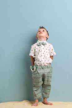 little boy in fashionable clothing and a slingshot in his hands stands on the sand on a light blue background