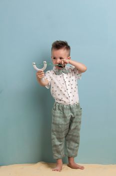 little boy in fashionable clothing and a slingshot in his hands stands on the sand on a light blue background