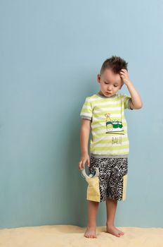 little boy in shorts and a T-shirt with a slingshot in his hands stands on the sand on a light blue background BALIQ - A FISH