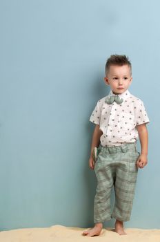 little boy in fashionable clothing and a slingshot in his hands stands on the sand on a light blue background