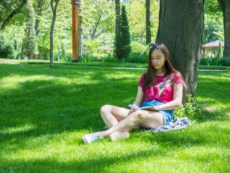 young girl reading a book in a picturesque park, sunny day. the girl of Asian appearance