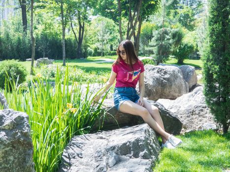 a young girl is sitting on a large rock in a picturesque park in sunny day. the girl of Asian appearance