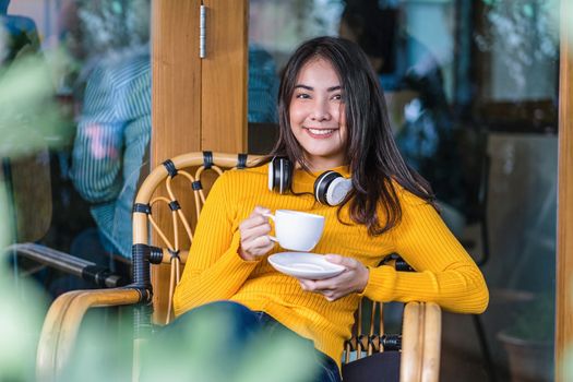 Asian young female holding a cup of coffee and sitting in modern coffee shop or coworking space beside window mirror, wearing Headphones for listening the music,hipster lifestyle and freelance concept