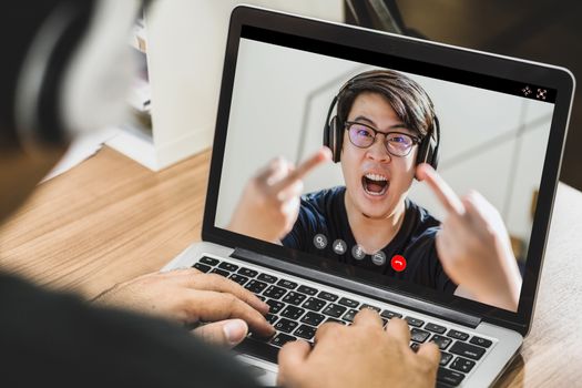 Asian business man wearing headphone and using laptop for video conference with angry colleague showing thumbs up sign Fuck when online meeting, doing fucking off to camera, Social distancing concept