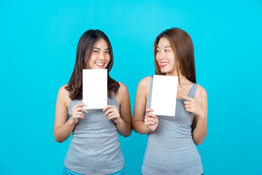 Two Asian smiling young women showing and presenting with posters on isolated blue color background, wearing vest in summer season indoors studio, Brochure and white blank paper for advertisement