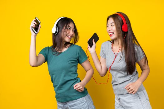 Two Happiness Asian smiling young woman wearing wireless headphones for listening music via smart mobile phone and dancing on isolated yellow color background, Lifestyle and leisure with hobby concept