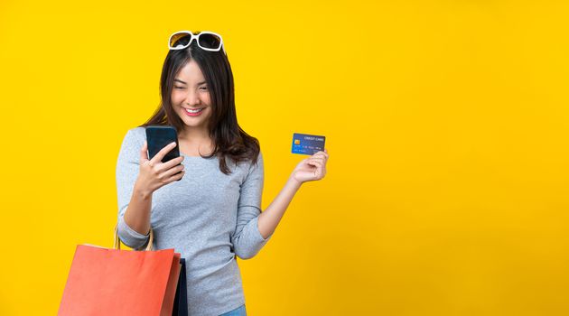 Happy Asian smiling woman using mobile phone and credit card and Carrying a shopping colorful bag for online shopping on isolated yellow color background, copy space and studio concept