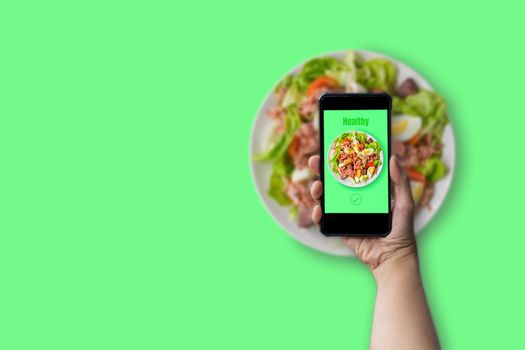 Healthy food concept. Hand holding smartphone with green text healthy with tuna salad, boil egg, lettuce and tomato on white plate on green background with copy space. 