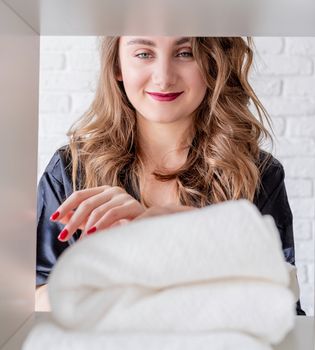 Laundry concept. Smiling young woman arranging a pile of towels on a shelf on white bricks background