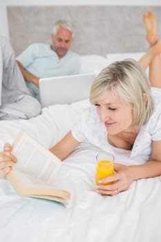 Closeup of a woman reading book with man using laptop in background in bed at home
