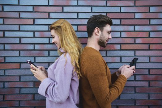 Side portrait of beautiful young couple is using smartphones and smiling, standing back to back against brown brick wall.