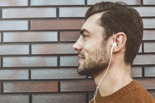 Photo of stylish handsome young man with bristle standing outdoors. Man wearing brown sweater. Smiling man listening to music on headphones and leaning against brick wall.