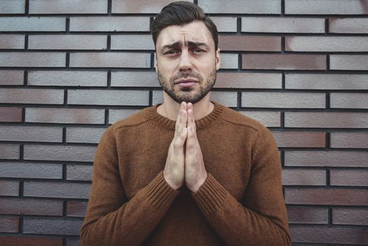 Young adult man standing over white brick wall begging and praying with hands together with hope expression on face very emotional and worried.
