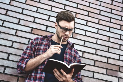 Young hipster guy wearing eyeglasses reading a book or note book make some notes and ideas on brick wall background.
