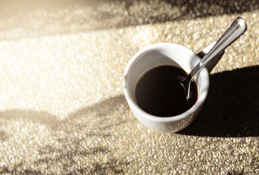 a white ceramic cup with a steel spoon filled with black coffee illuminated by natural sunlight. Coffee break. Caffeine and energy drinks. Quiet scene