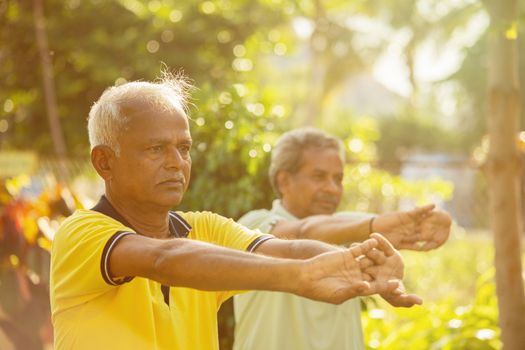 Two old men stretching hands, doing yoga at park with backlight contrast - concept of active, healthy and fitness of seniors