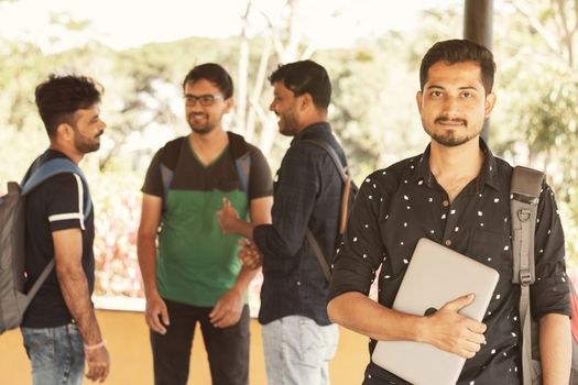 Smiling young adult student with holding laptop in hand while friends staning in background - Concfident graduate scholar smiling at university college