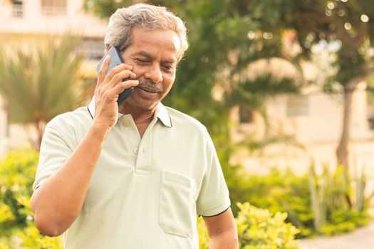 Healthy senior Asian on phone outdoor green park - Elder man busy in mobile - Happy male parent talking on phone