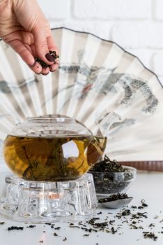 Glass teapot with green tea and chinese fan background. Hand putting tea into the pot