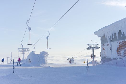 two skiers head down a run having disembarked from ski lifts that rise up above the clouds into the pale blue sky. Very cold and snowy conditions.