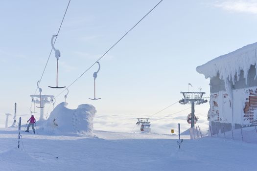 a single skier heads down a run having disembarked from ski lifts that rise up above the clouds into the pale blue sky. Very cold and snowy conditions.
