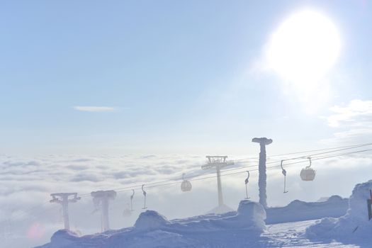 The sun flares from behind these gondola and T-bar ski lifts as they rise, frozen in snow and ice, from beneath the rolling clouds. Harsh and cold.