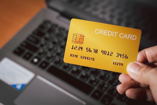 Shopping online and new normal concept. Hand holding yellow of credit card with blurry laptop background. Pay and buy online at home. 