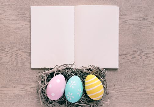 Pastel and colorful easter eggs on nest with blank notebook on wooden background, happy easter holiday concept