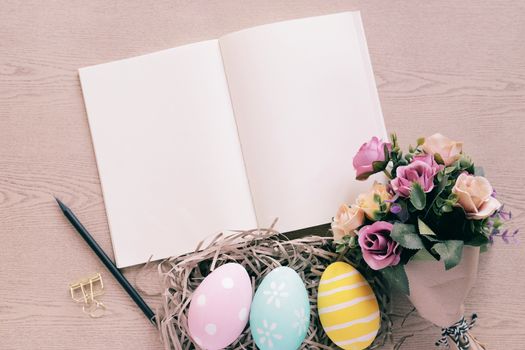 Pastel and colorful easter eggs in nest and bouquet of flower with blank notebook on wooden background, happy easter holiday concept