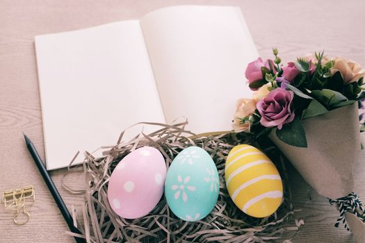 Pastel and colorful easter eggs in nest and bouquet of flower with blank notebook on wooden background, happy easter holiday concept