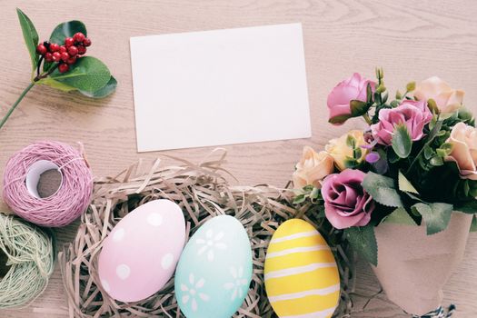 Pastel and colorful easter eggs in nest and bouquet of flower with blank card on wooden background, happy easter holiday concept