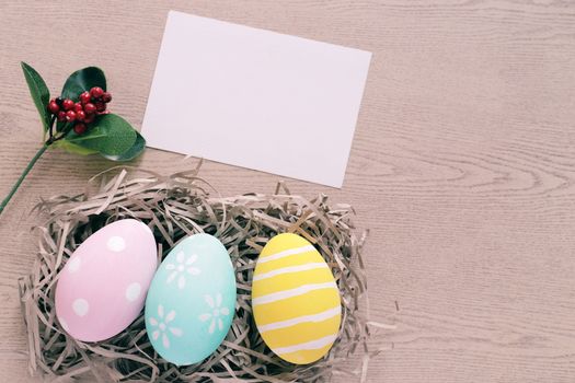 Pastel and colorful easter eggs in nest with blank card on wooden background, happy easter holiday concept