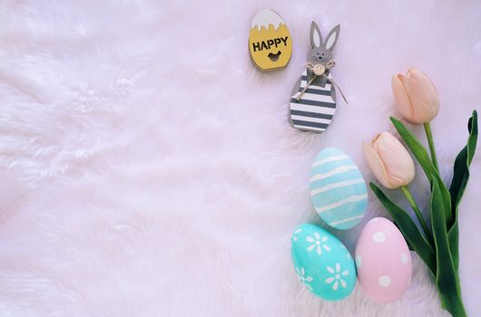 Happy Easter concept with wooden bunny and colorful easter eggs on white fur background and pink tulips. Top view with copy space 