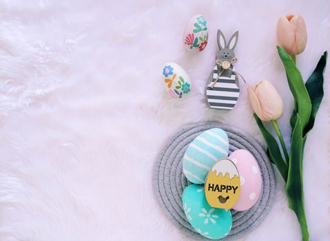 Happy Easter concept with wooden bunny and colorful easter eggs on white fur background and pink tulips. Top view with copy space
