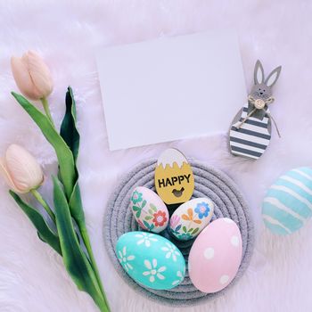 Happy Easter concept with wooden bunny and colorful easter eggs on white fur background and pink tulips. Top view 