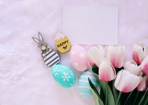Happy Easter concept with wooden bunny and colorful easter eggs on white fur background and pink tulips. Top view with copy space 