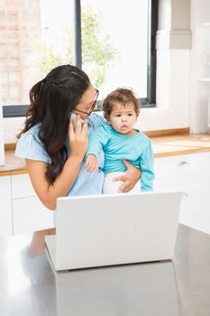 Smiling brunette holding her baby and using laptop on phone call in the kitchen