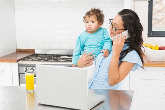 Smiling brunette holding her baby and using laptop on phone call in the kitchen