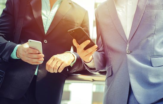 Midsection of two colleague businessmen using smartphone at the outside of modern office building, business and technology concept