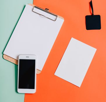 Creative flat lay of smartphone, blank clipboard and white card on minimal color background