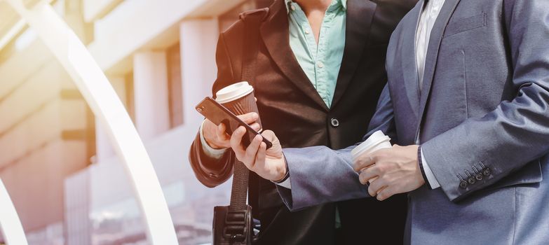 Midsection of two colleague businessmen using smartphone and holding coffee cup at the outside of modern office building with copy space and sunlight tone