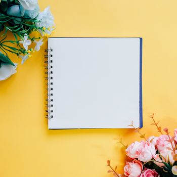 Flat lay of minimal workspace blank notebook with flower on yellow background, spring and summer concept