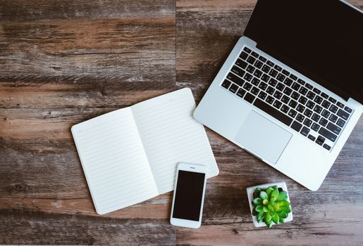 Flat lay photo of workspace desk with laptop, smartphone, blank notebook and green plant with copy space wooden background