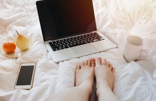 Close up Woman feet relaxing on bed with a laptop computer, smartphone, tea and fruits on white bed sheet