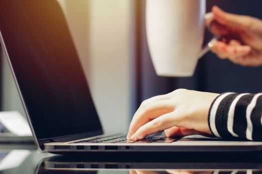 Female hands typing on keyboard of laptop while holding cup of coffee, working at home concept