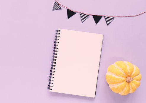 Flat lay of minimal workspace blank notebook with pumpkin and bunting flag on purple background, copy space