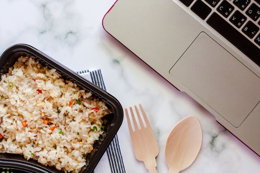 Delicious fried rice in lunch box on napery with wooden spoon and fork and computer laptop at workplace for ready to eat and food concept