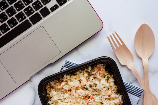 Delicious fried rice in lunch box on napery with wooden spoon and fork and computer laptop at workplace for ready to eat and food concept
