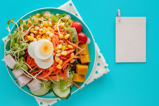 Fresh salad vegetable with boiled chicken egg and blank note paper on blue background for healthy eating and diet food concept