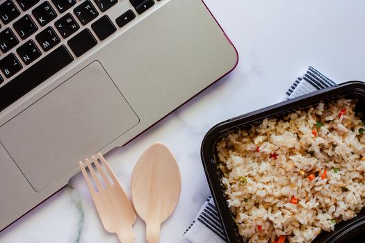 Delicious fried rice in lunch box on napery with wooden spoon and fork and computer laptop at workplace for ready to eat and food concept
